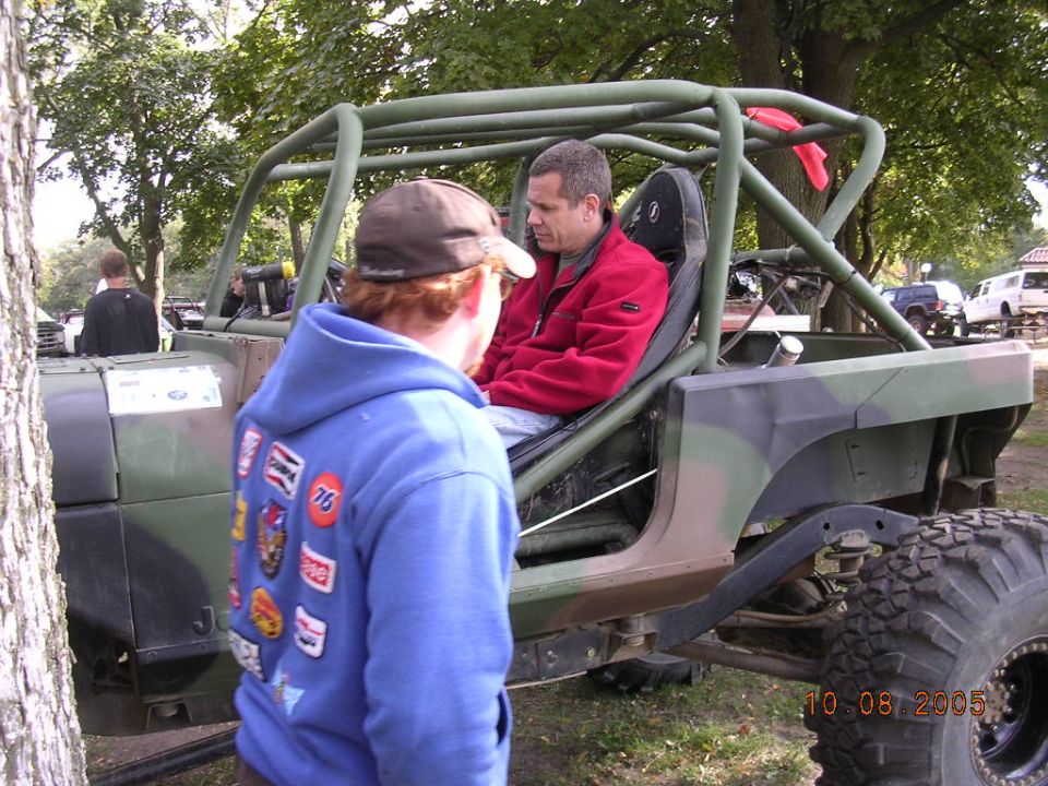 Modified Jeep CJ-7 buggy with roll cage and camouflage paint, two men inside. Another Jeep and a white truck are in the background