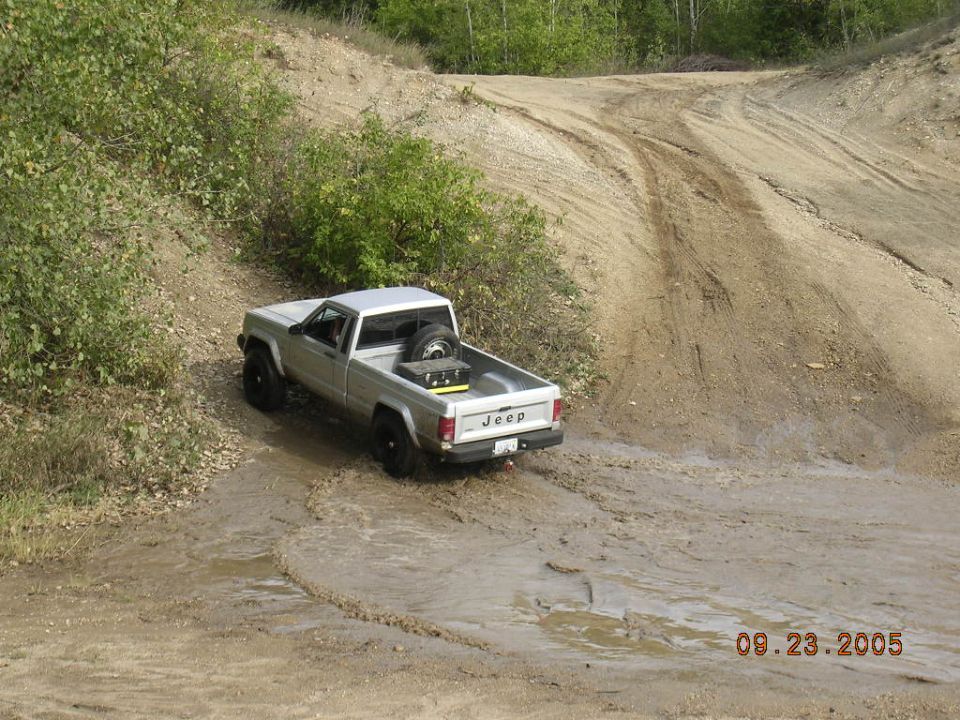 Silver Jeep Comanche pickup truck with spare tire and toolbox in bed, driving through mud on a dirt track. Date stamp reads 09.23.2005