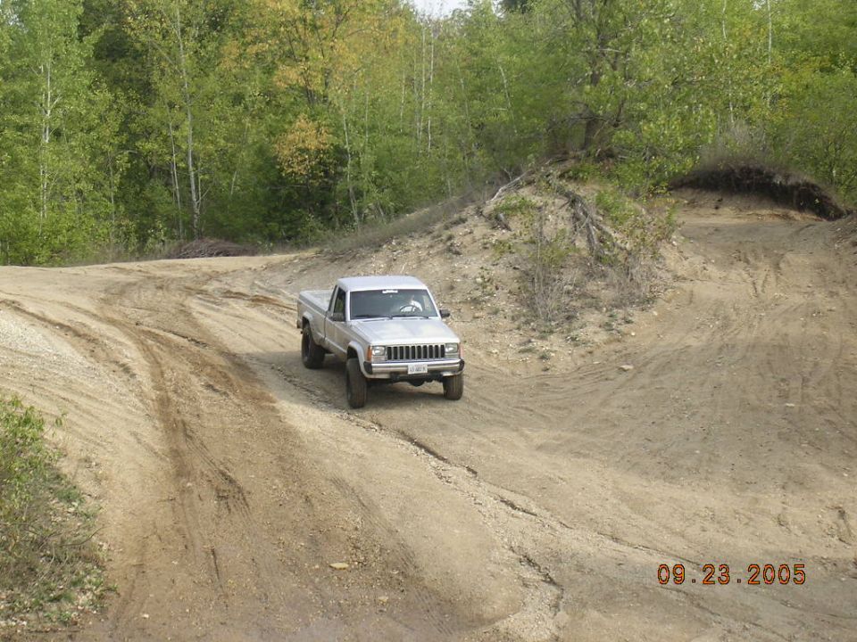 Silver Jeep Comanche pickup truck driving on a dirt road with trees in the background. The date 09.23.2005 is visible
