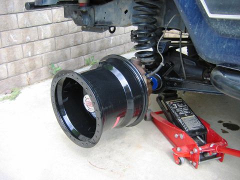 1985 Ford Bronco - The suspension and wheel assembly of a blue vehicle, likely a Ford Bronco, is shown with a red jack supporting it.
