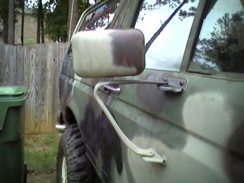 1988 Ford Bronco - A side view of a camouflage-painted SUV, focusing on the side mirror and door, with a trash can and fence visible.