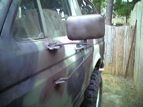 1988 Ford Bronco - This camouflage-painted SUV is seen from the side, a large side mirror and a wooden fence in the background.