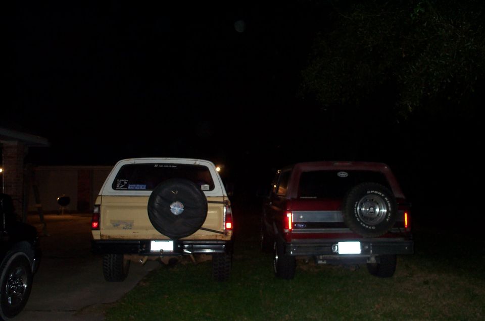 Two SUVs parked at night, rear view. The one on the left is tan with a spare tire on the back. The one on the right is red with a spare tire on the back