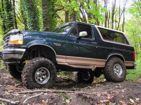 1995 Ford Bronco - A lifted 1995 Ford Bronco navigates a muddy trail surrounded by trees.