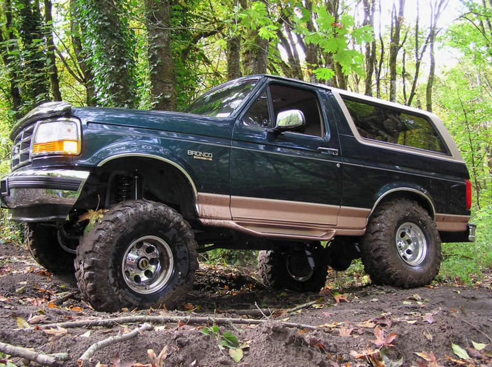 Lifted green and tan Ford Bronco on mud-terrain tires parked on a muddy forest trail