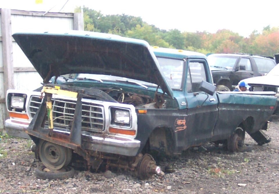 Front end of a green and black Ford F-Series truck with hood open, missing front wheels, and a large metal contraption attached to the bumper