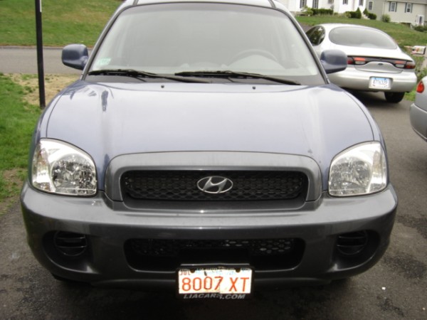 Front view of a blue Hyundai Santa Fe with a dark gray grille and bumper. A silver Ford Taurus is parked behind it