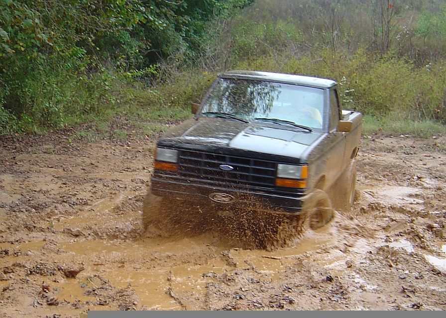 Black pickup truck with a dented front fender driving through deep mud, splashing water