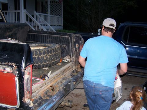 1979 Ford F-150 - A man loads items into the bed of a worn pickup truck, with a spare tire inside.