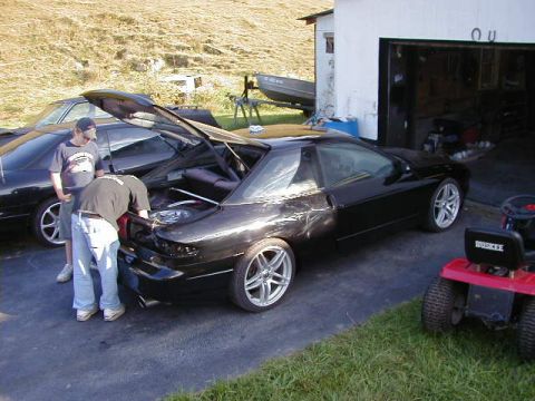 1994 Ford Probe - Two black coupes are parked outside a garage, with people attending to the open trunk of one car.