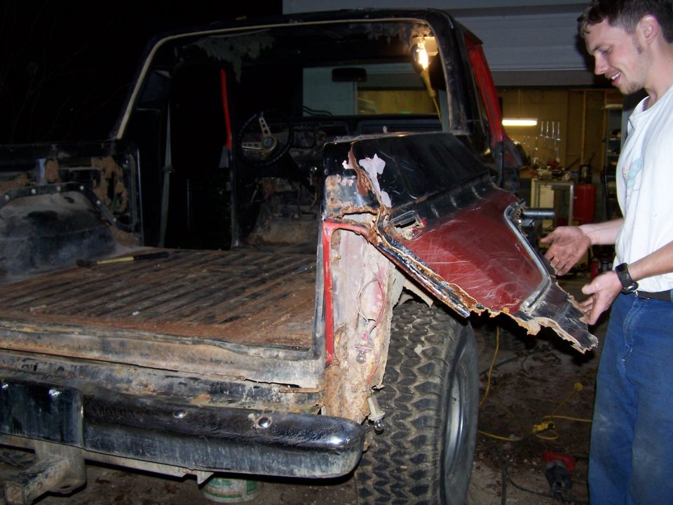 Heavily rusted red and black pickup truck bed and rear quarter panel, with a man pointing at the damage