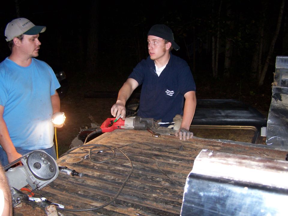 Two men work on a dark, wooden truck bed at night with tools. One man holds a flashlight