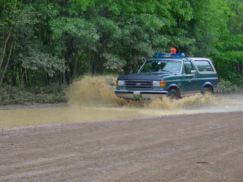 1989 Ford Bronco - 1989 Ford Bronco navigates a muddy obstacle on a dirt path surrounded by trees.