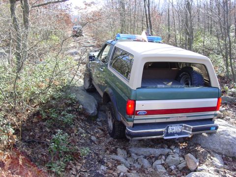 1989 Ford Bronco - A 1989 Ford Bronco navigates a challenging, rocky off-road path with another vehicle in the distance.