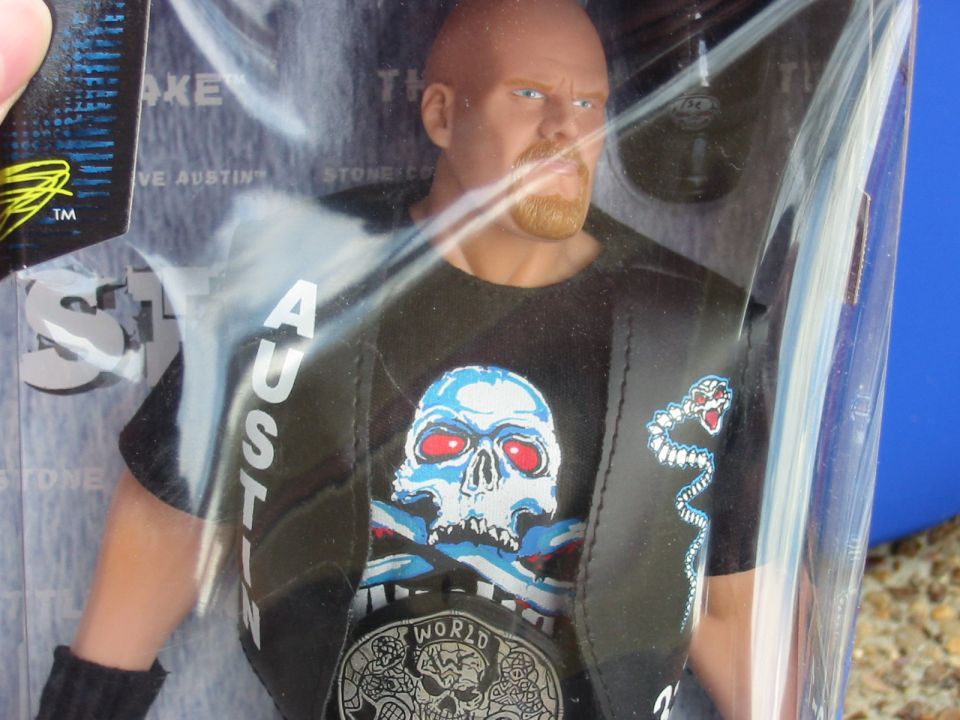 Action figure of Stone Cold Steve Austin in packaging, wearing a black shirt with a skull graphic and a championship belt