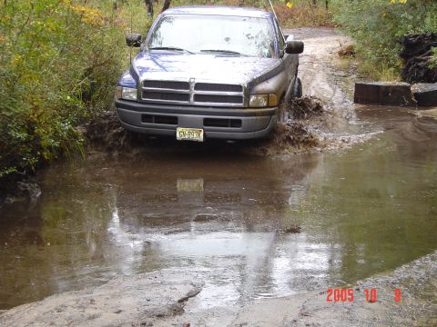 2005 Dodge 1500 - A blue pickup truck navigates through a water-filled path, kicking up spray.