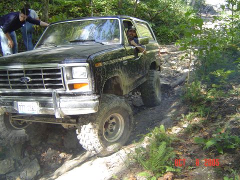 1986 Ford Bronco - A black 1986 Ford Bronco tackles a challenging off-road course with two people visible inside and two outside.