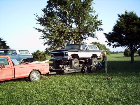 1986 Ford Bronco - A lifted Ford Bronco is transported on a trailer towed by a red pickup truck, with another SUV in the background.