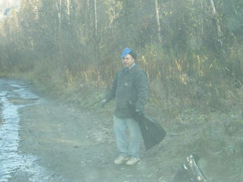2000 Ford E350 - A man stands on a dirt road holding a black trash bag, with a wooded area behind him.