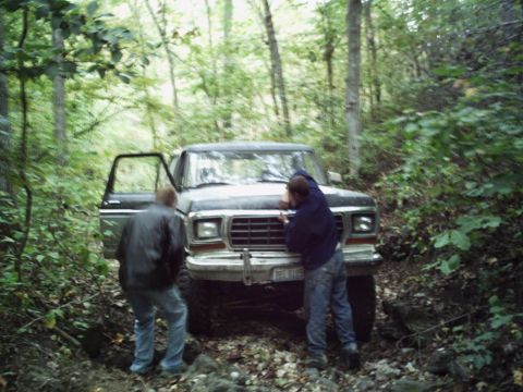 1986 Ford Bronco - A Ford F-Series truck is stopped on a rough, wooded path with two people examining the front.