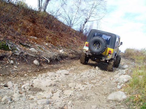 2000 Ford E350 - A yellow Jeep Wrangler navigates a challenging, rocky off-road path surrounded by trees and dry foliage.