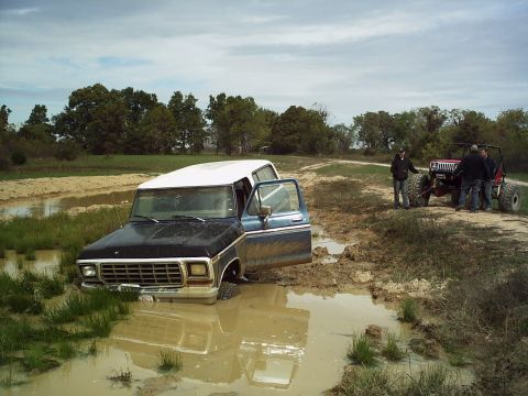1986 Ford Bronco - A blue and white Ford Bronco is stuck in a muddy field with its door open, while another off-road vehicle stands nearby.