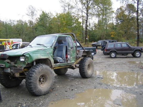 1990 Ford Bronco - Heavily modified green Toyota pickup truck with oversized tires and a custom roll cage sits in a muddy area, with other vehicles visible in…