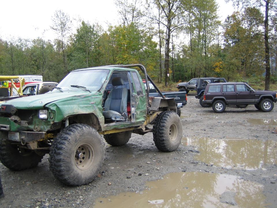 Modified green Toyota pickup truck with large tires and roll cage, parked in a muddy lot with other vehicles