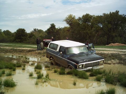 1986 Ford Bronco - A dark blue and white SUV is stuck in a muddy field, with a red Jeep and people in the background.