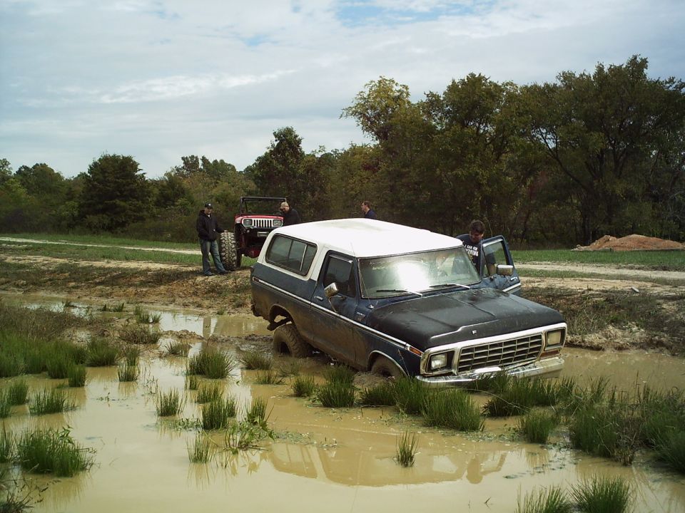Dark blue and white SUV stuck in mud, front end submerged. A red Jeep is behind it with two people standing nearby
