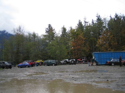1990 Ford Bronco - A collection of off-road vehicles are gathered in a muddy area, with a large blue container and trees in the background.