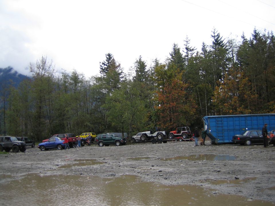 Several off-road vehicles parked in a muddy lot with trees and a blue shipping container