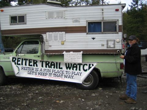 1990 Ford Bronco - A woman stands next to a pickup truck with a camper and a large banner promoting Reiter Trail Watch.
