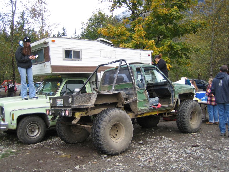 Modified green truck with large tires and roll cage, parked next to a pickup with a camper shell. People are standing around