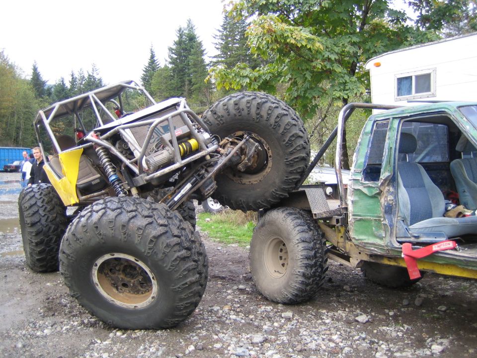 Two heavily modified off-road vehicles, one yellow with a roll cage, the other a green pickup truck, are positioned closely together on a dirt surface
