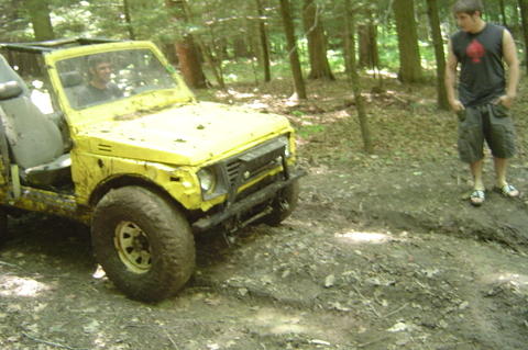 1987 Suzuki Samurai - A yellow 1987 Suzuki Samurai navigates a muddy trail in a wooded area, with a person standing nearby.