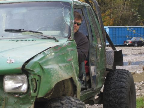 1990 Ford Bronco - A man sits in a heavily damaged green off-road vehicle, with another car visible in the background.