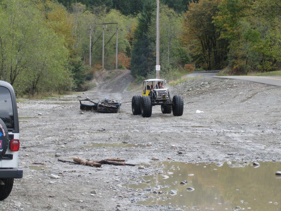 Yellow buggy with big tires tows a pile of debris on a dirt road. White Jeep visible on left
