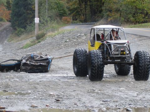 1990 Ford Bronco - A custom off-road vehicle tows a mangled car chassis across a muddy, gravelly surface.