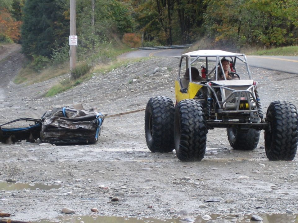 Yellow off-road buggy with large tires tows a crushed car on a dirt road