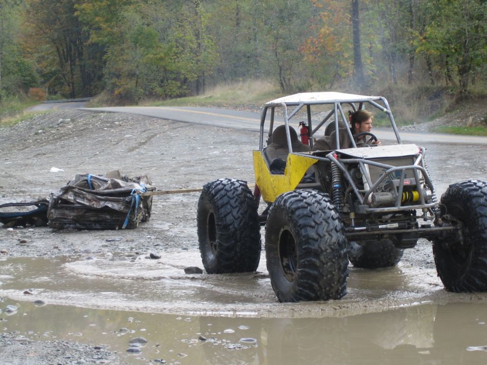 Yellow off-road buggy with roll cage and large tires tows a pile of scrap metal through a muddy puddle