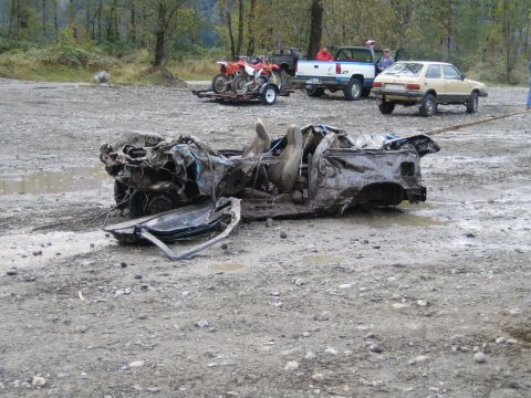 1990 Ford Bronco - A heavily damaged car chassis lies on the ground, with other vehicles and a trailer visible in the background.