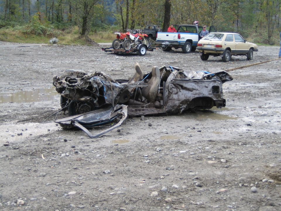 Wrecked car chassis with open doors and exposed interior, resting on a gravelly, muddy ground