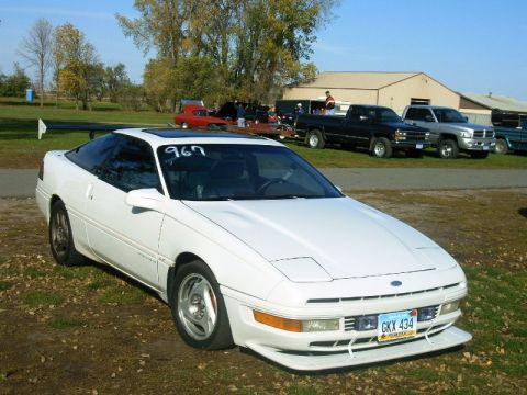 1992 Ford Probe - A white 1992 Ford Probe, modified with a large rear spoiler and racing numbers, sits parked at an outdoor event.