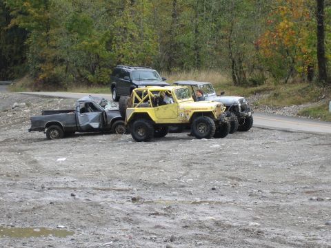 1990 Ford Bronco - Multiple off-road vehicles and a damaged pickup truck are parked on a gravel lot with trees in the background.