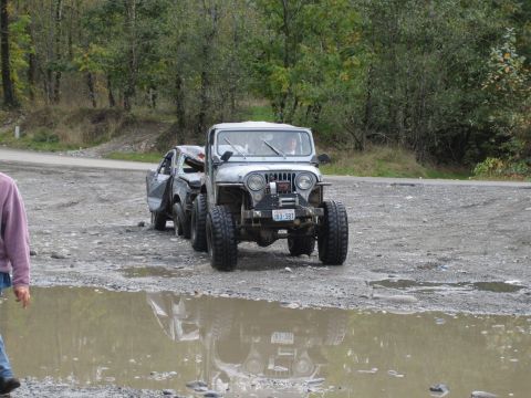 1990 Ford Bronco - A modified Jeep tows a damaged car through a muddy, gravelly area with trees lining the background.