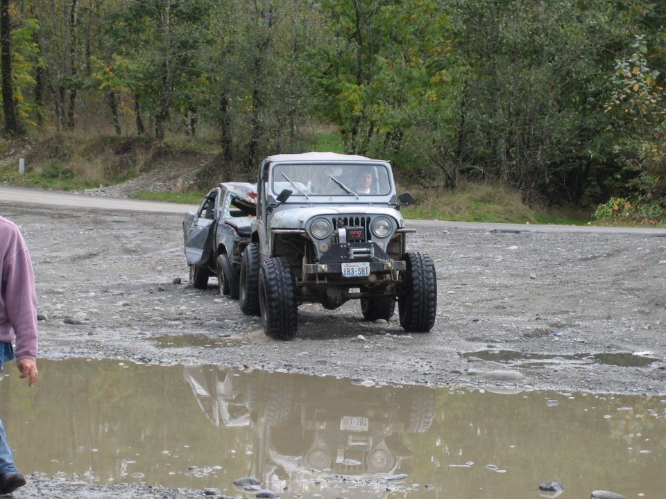 Jeep towing a beat-up car on a dirt road with trees in background. Puddle in foreground reflects the Jeep