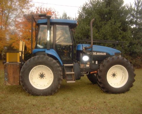 1985 Ford F-150 - Side view of a blue New Holland tractor parked on grass with trees in the background.