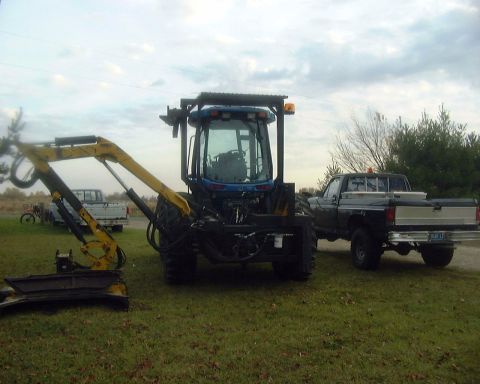 1985 Ford F-150 - A pickup truck and a tractor with an excavator attachment are parked on a grassy area under a cloudy sky.