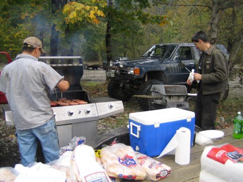 1990 Ford Bronco - A dark blue SUV with significant off-road modifications is parked at a campsite where people are grilling.